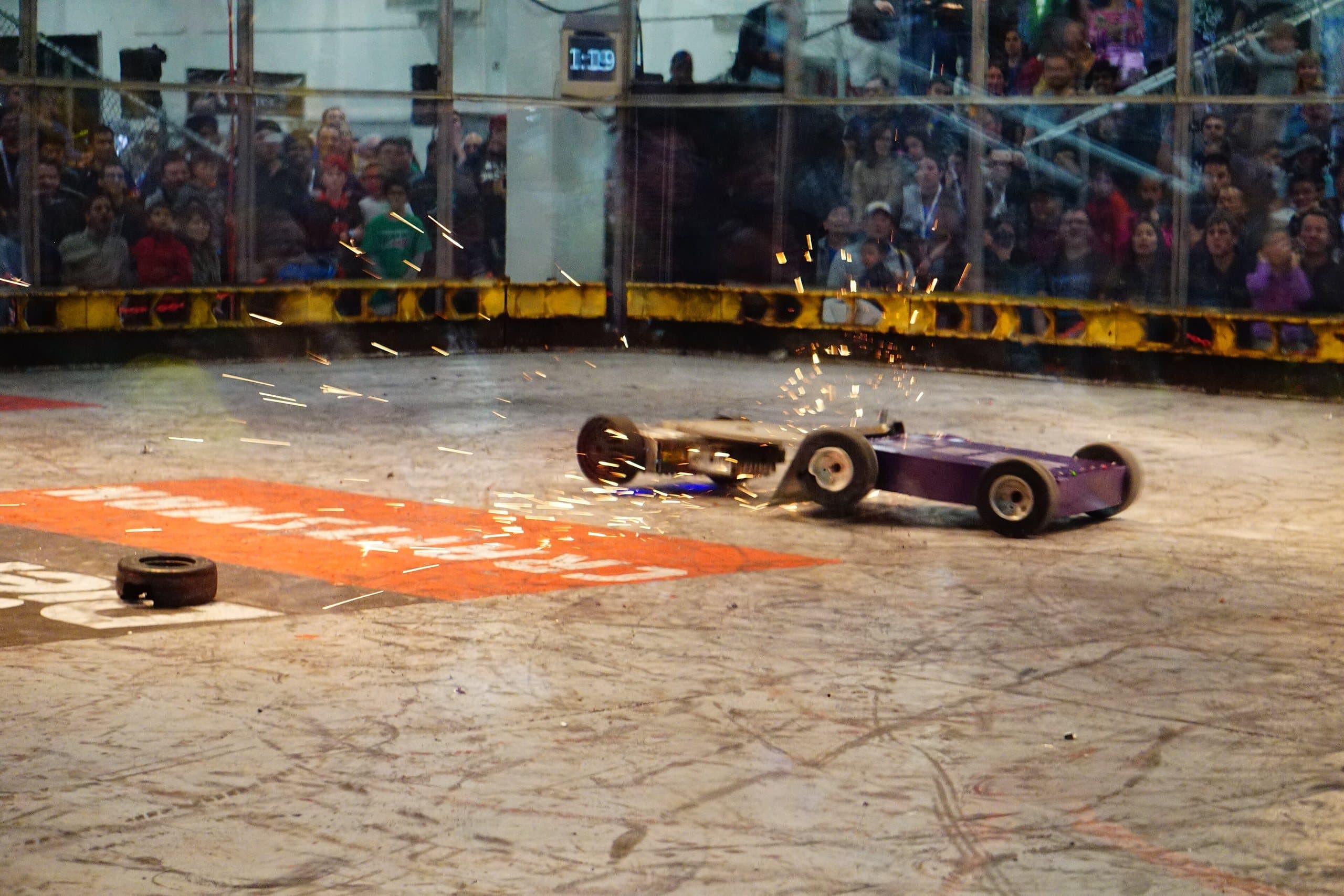 Robôs de rodinhas em combate numa arena de batalha, com faíscas e público